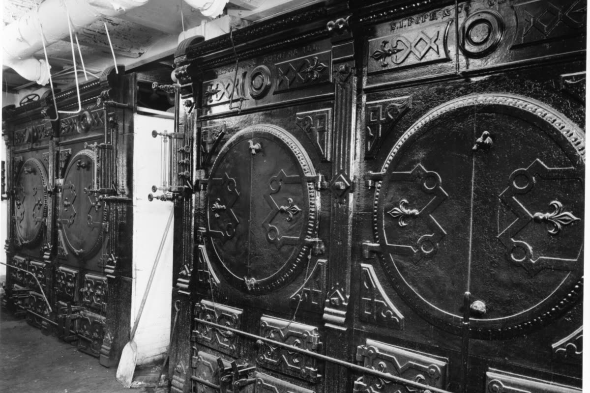 The ornate cast-iron doors of an old basement furnace in a Rochester, New York house, with pipes overhead and a shovel leaning beside them